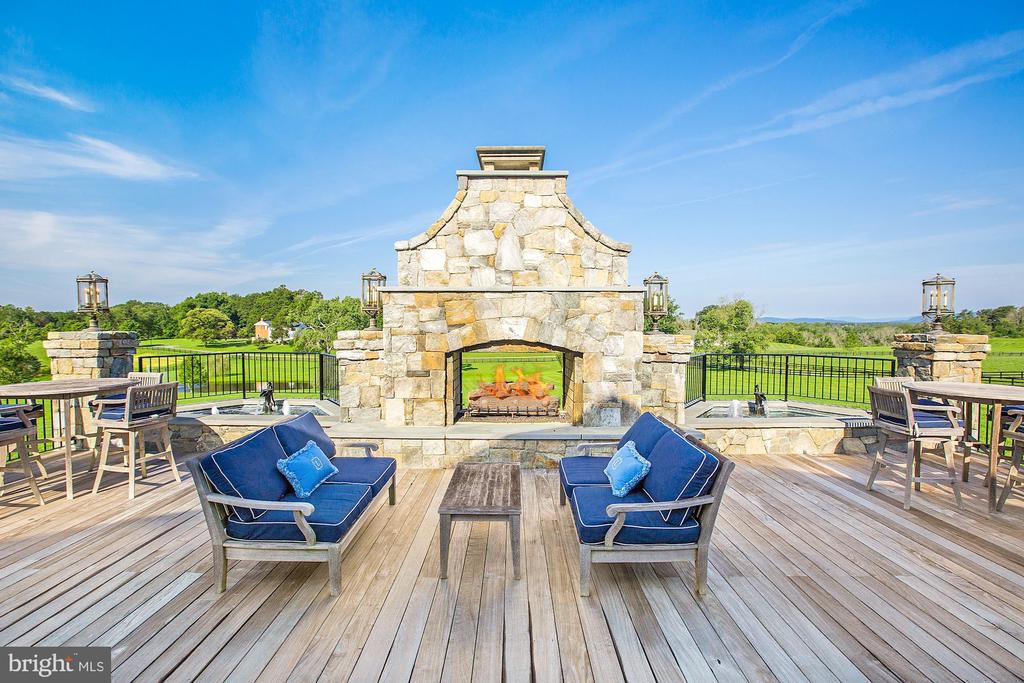 Outdoor deck with a stone fireplace, blue-cushioned seating, and views of green fields.