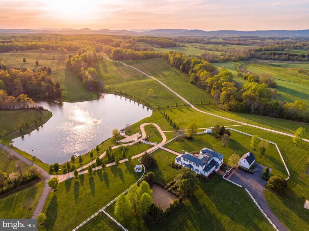 Country estate with a large white house, pond, landscaped grounds, and fields at sunset.