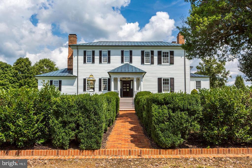 White two-story house with dark shutters, brick chimneys, and a brick walkway through clipped hedges.