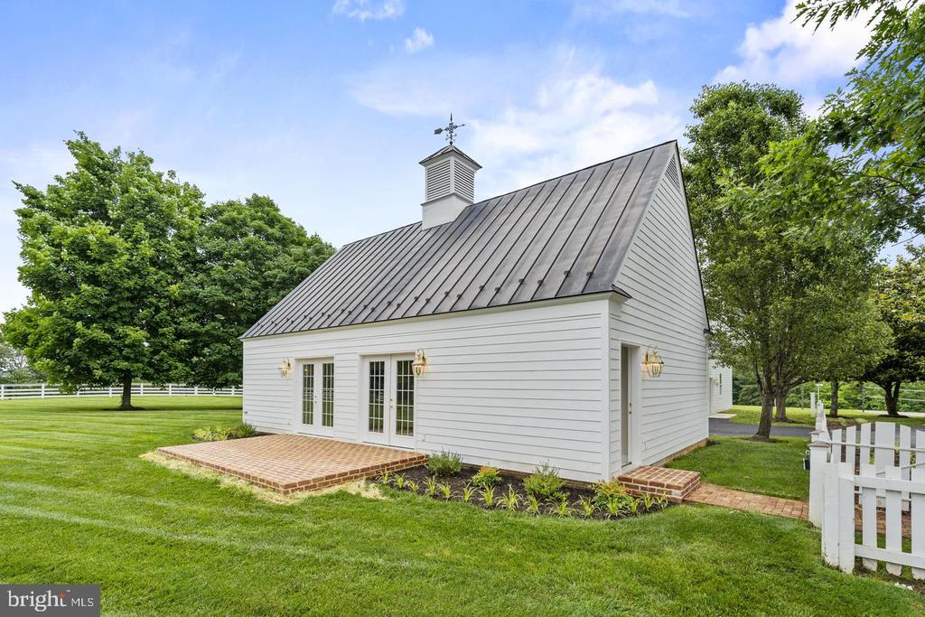 White detached building with a metal roof, cupola, brick patio, and white fencing.