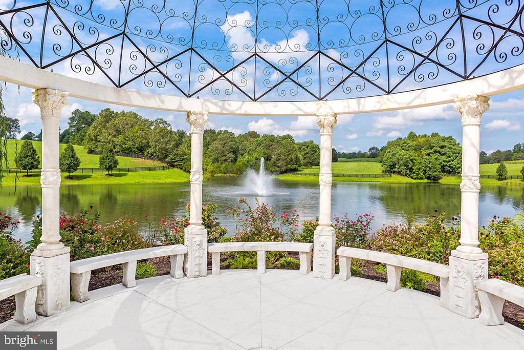 White-columned gazebo overlooking a pond with a fountain and rolling green grounds.
