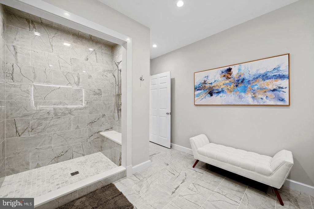 Bathroom with a marble walk-in shower and white chaise lounge beneath abstract artwork.