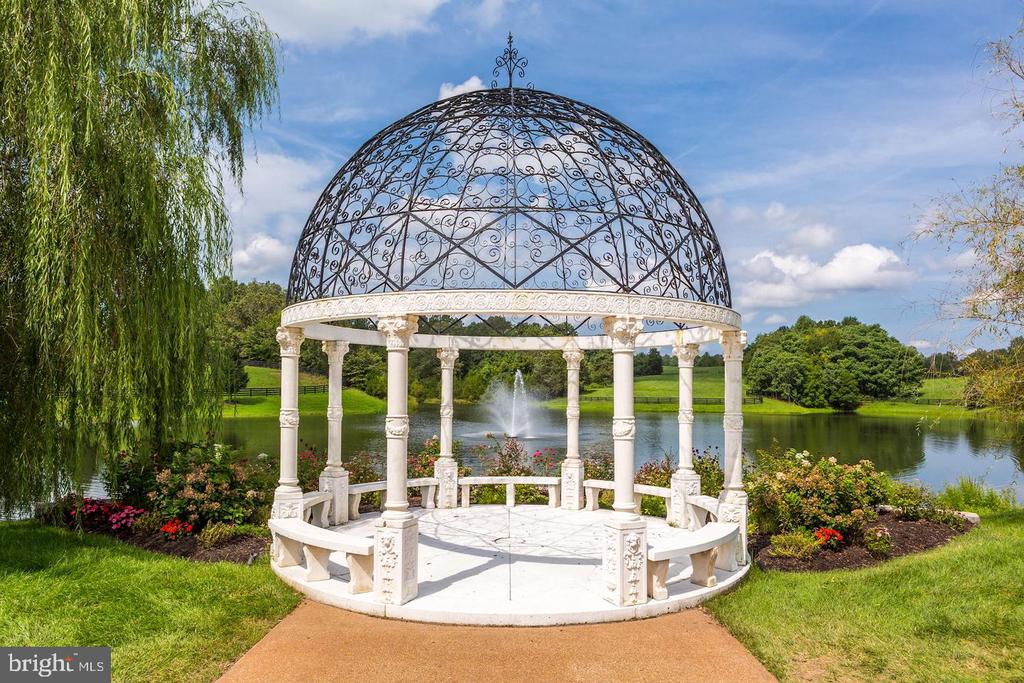 Ornate white gazebo beside a pond with a fountain and landscaped grounds.