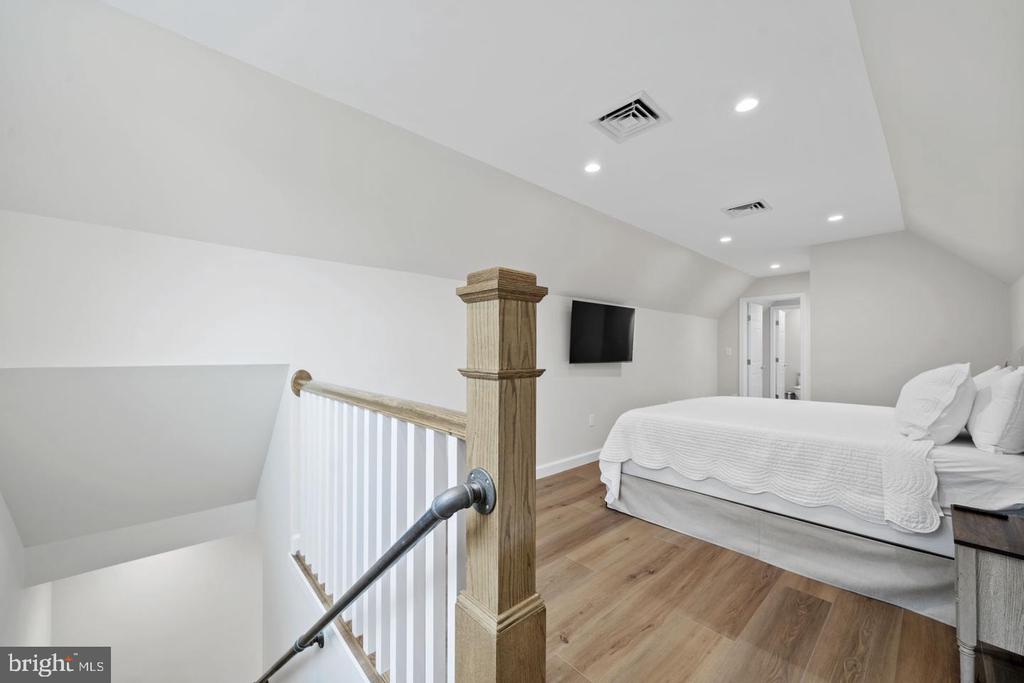 Upstairs bedroom with a white bed, light wood floors, and a wooden railing beside the stairwell.