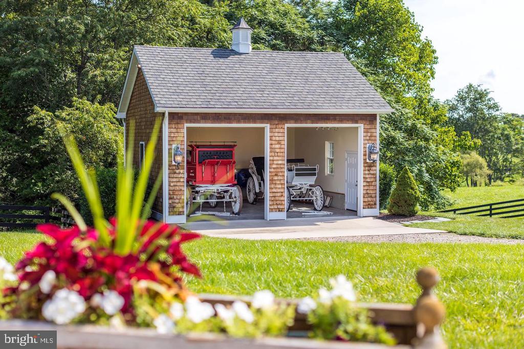 Detached carriage house with two open bays displaying horse-drawn carriages, surrounded by trees and lawn.