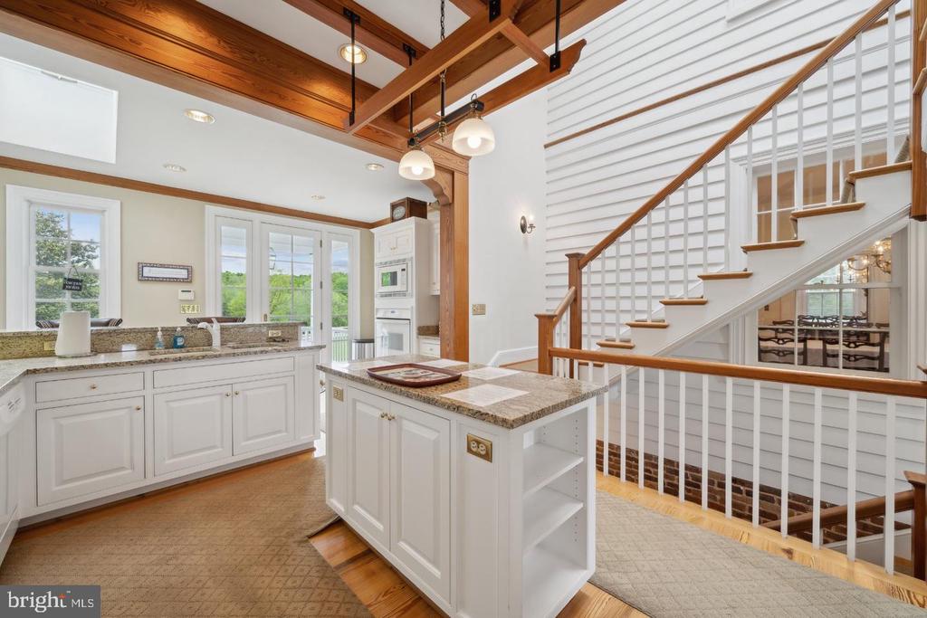 Open kitchen with granite island, white cabinetry, exposed timber beams, and a prominent staircase.