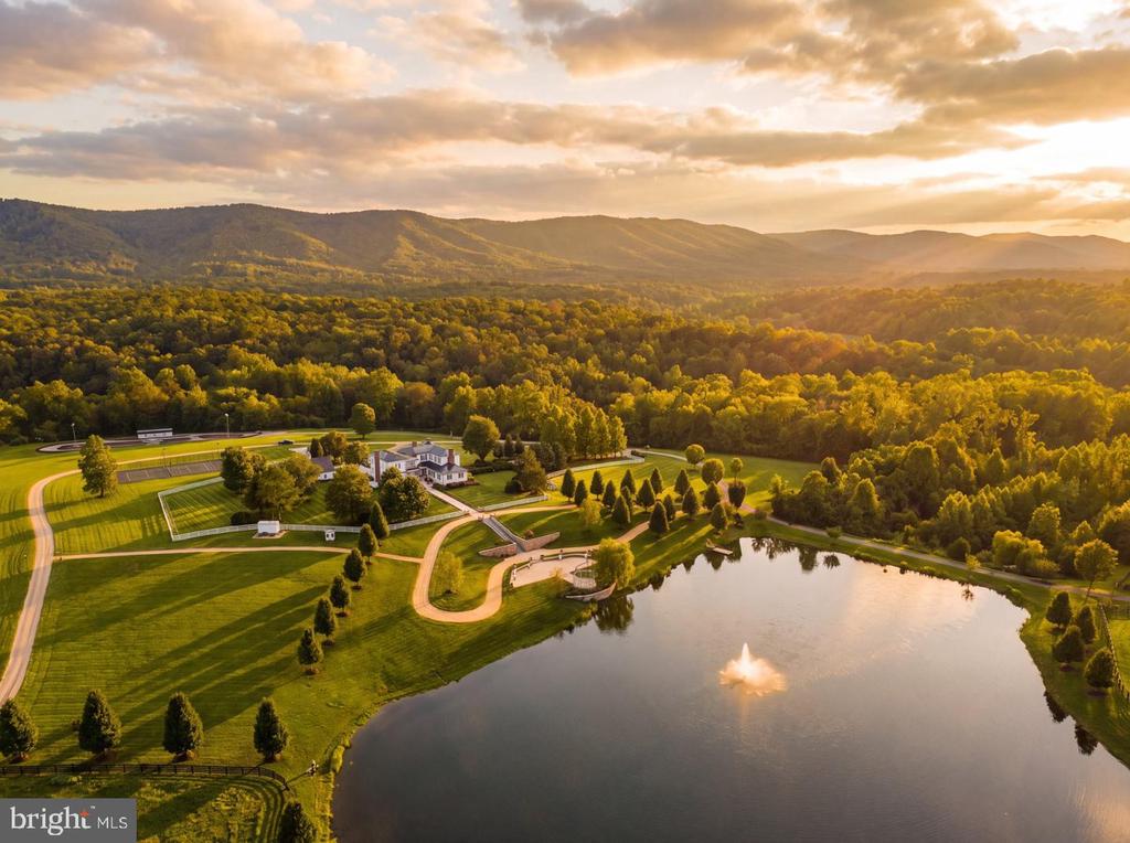 Aerial view of a landscaped estate with a pond, sports courts, wooded hills, and distant mountains.
