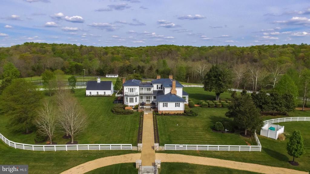 Aerial view of a white country home with a long driveway, white fencing, and wooded hills beyond.