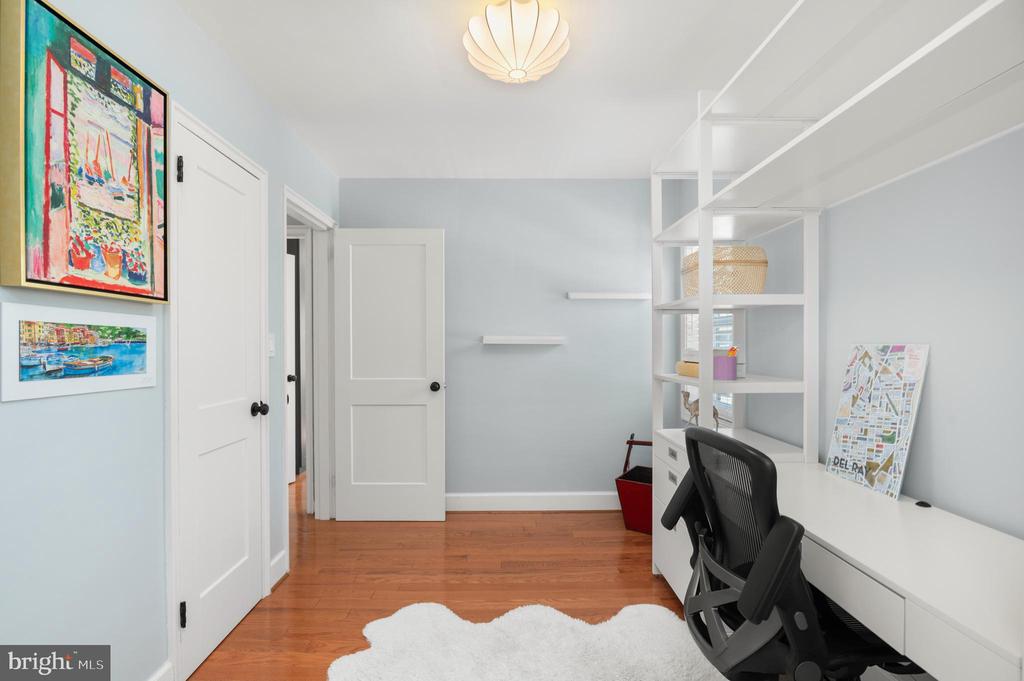 Home office with white built-in desk and shelving, black chair, hardwood floors, and colorful artwork.
