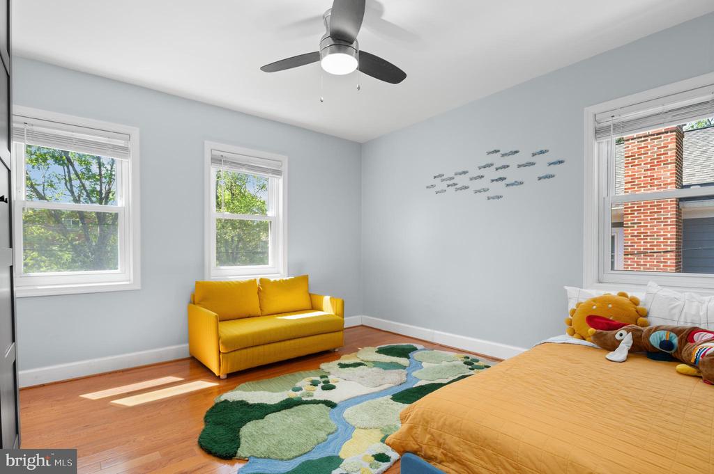 Bedroom with a yellow loveseat, orange-covered bed, and colorful rug beside sunlit windows.