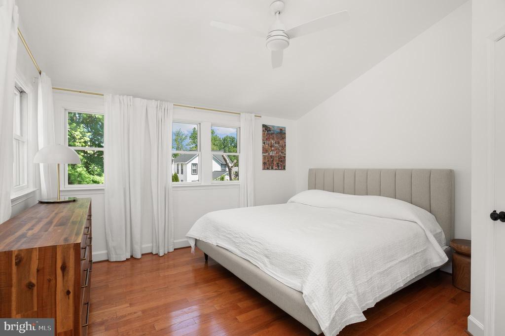 Bedroom with a white bed, gray upholstered headboard, hardwood floors, and windows overlooking trees.