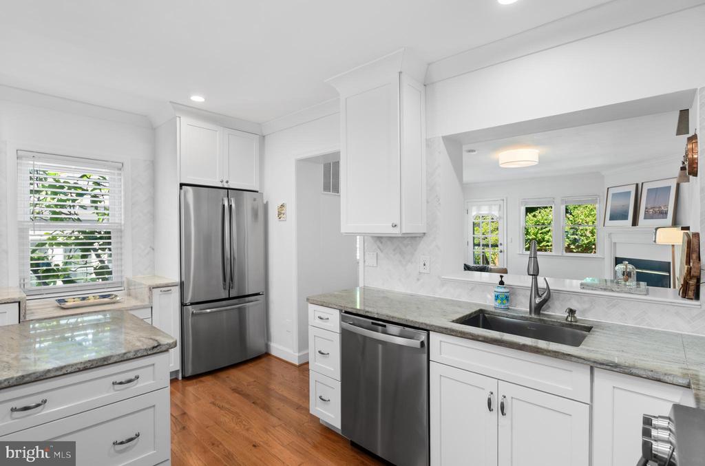 Renovated white kitchen with gray stone counters, stainless steel appliances, and a sink facing the adjoining living area.