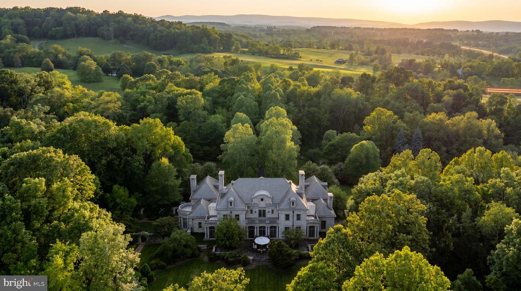Aerial view of a mansion nestled among wooded grounds, with rolling hills and fields beyond.