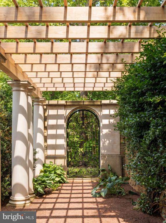Garden walkway beneath a wooden pergola, leading to an arched wrought-iron gate.
