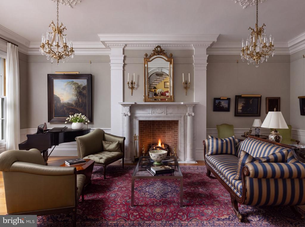 Living room with a lit fireplace, grand piano, chandeliers, and red patterned rug.
