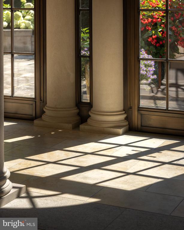 Sunlit interior with rounded columns, tiled floors, and tall windows overlooking flowering shrubs.