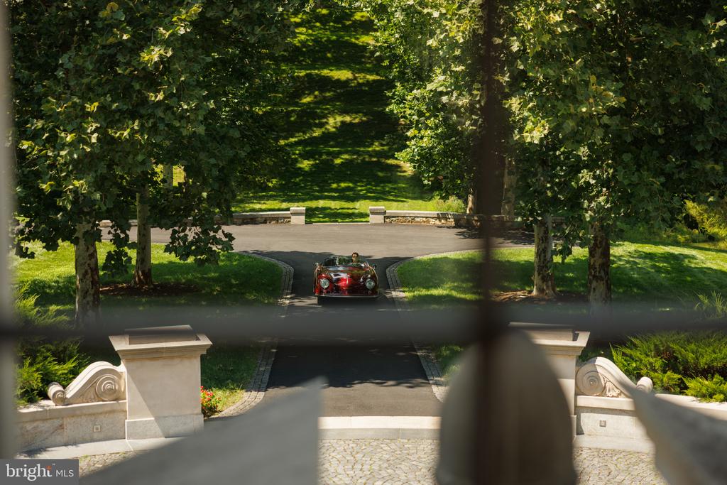 Tree-lined estate driveway framed by stone gateposts, with a vintage car approaching.