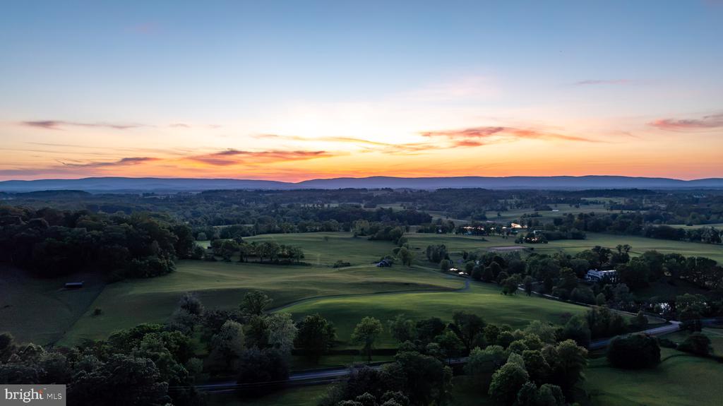 Aerial countryside at sunset, with rolling green fields, woods, and scattered homes.