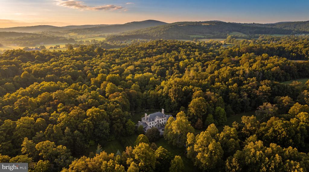 Aerial view of a large house among autumn woods, with rolling hills and distant mountains.