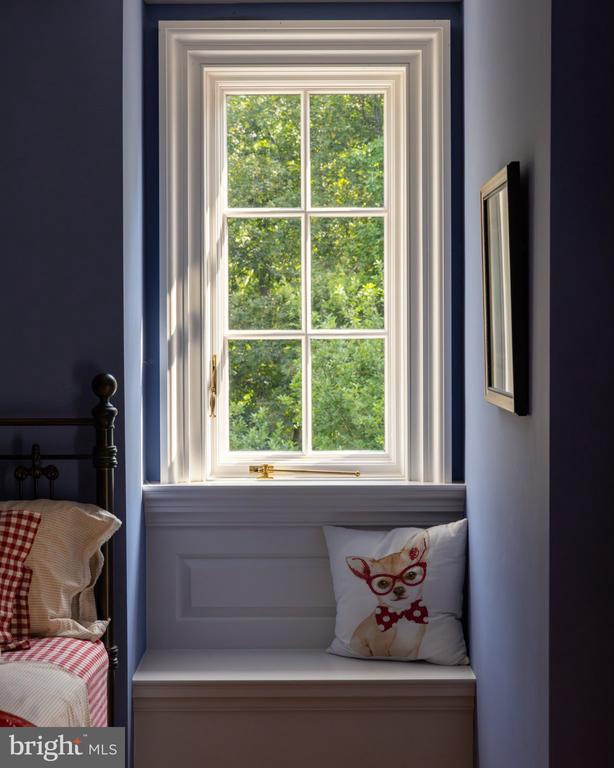 Bedroom window seat beneath a sunny, tree-facing window, with a cat pillow and gingham bedding.