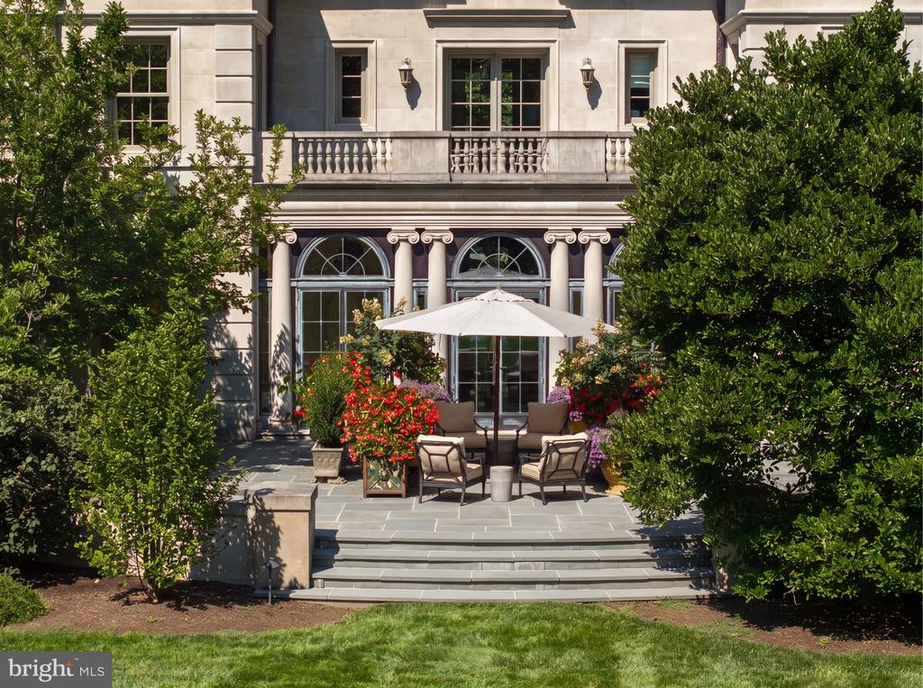 Stone terrace with umbrella-shaded seating and flower planters in front of an ornate home façade.