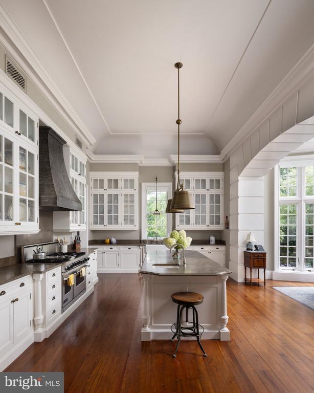 Kitchen with white cabinetry, a large stone-topped island, dark range hood, and warm wood floors.
