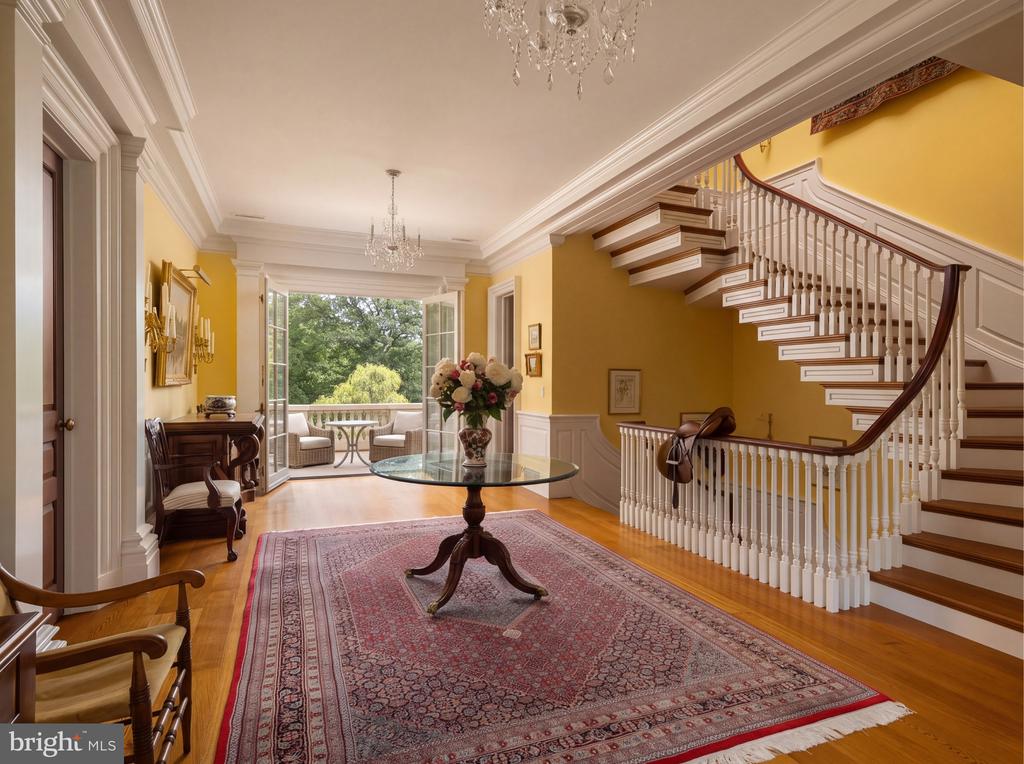 Furnished foyer with a sweeping staircase, patterned rug, and open doors overlooking a garden.