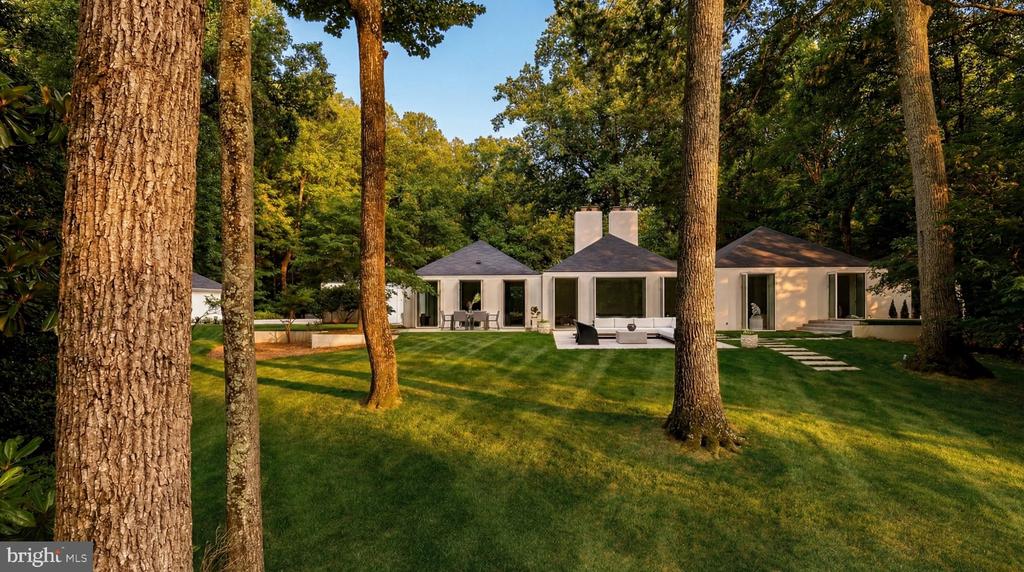 Modern home beyond a wooded lawn, with patio seating and a swimming pool.