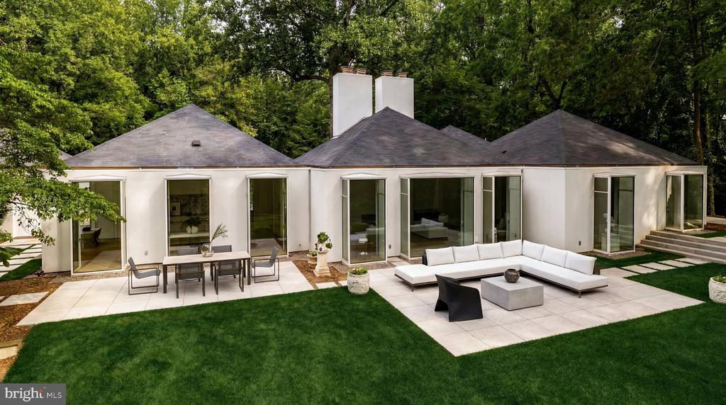 Modern white home with a broad stone patio, outdoor dining set, and white sectional beside a green lawn.