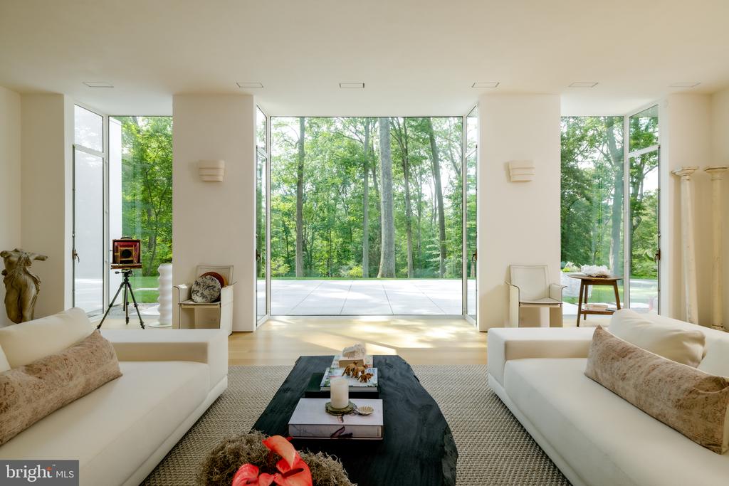 Living room with white sofas, floor-to-ceiling windows, and a leafy wooded view.