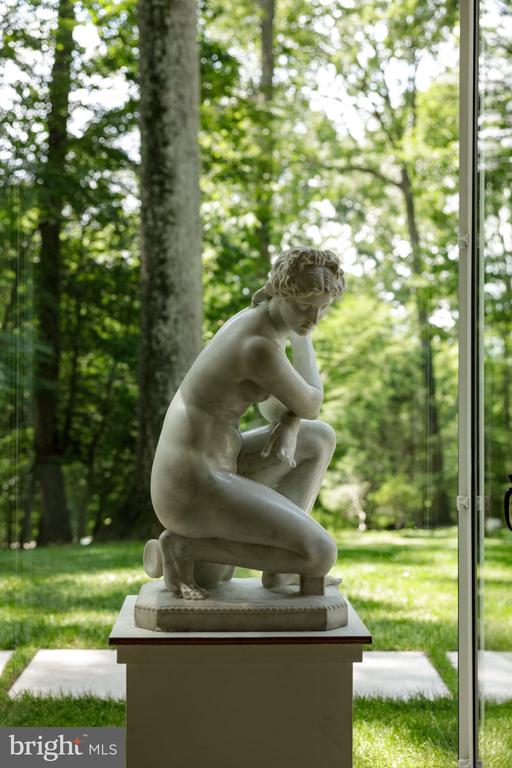Kneeling sculpture on a pedestal beside a glass wall overlooking wooded grounds.
