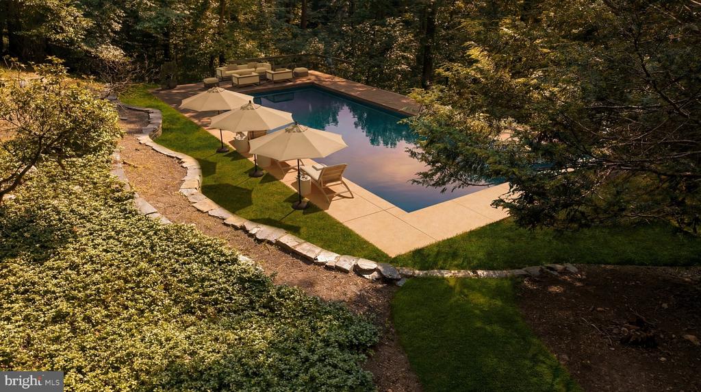 Swimming pool with a stone patio, lounge chairs, and umbrellas among mature trees.
