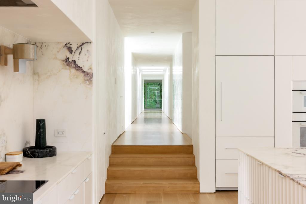Modern white kitchen with marble counters and island, opening onto a hallway with wooden steps.
