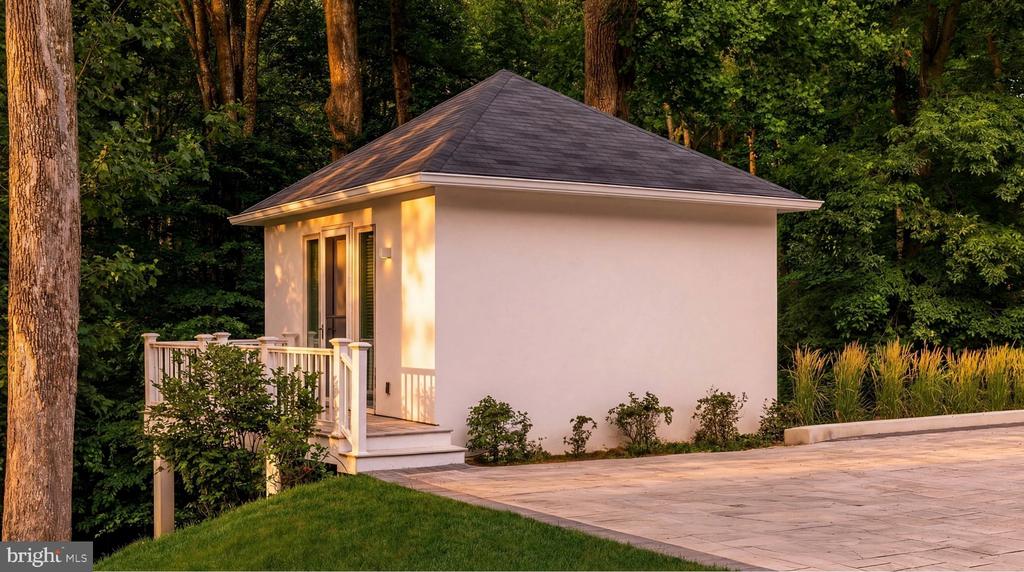 White detached outbuilding with a raised deck beside a stone patio, surrounded by mature trees.
