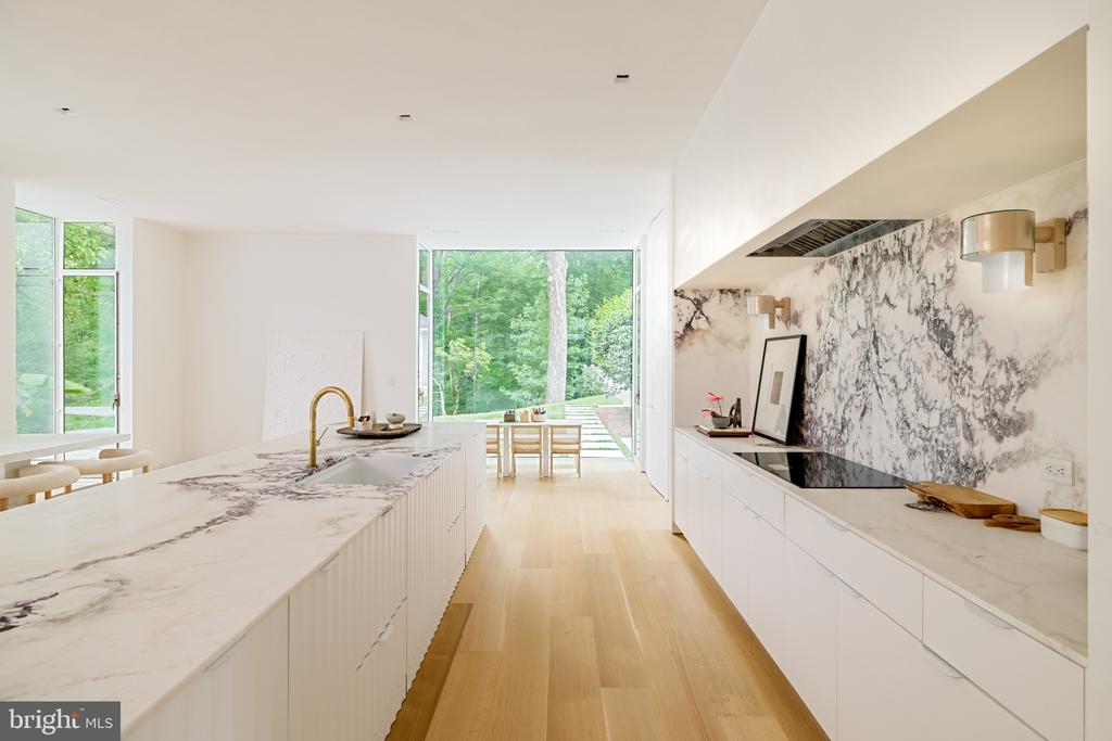 Modern kitchen with a marble island and backsplash, opening through large windows to a leafy garden.
