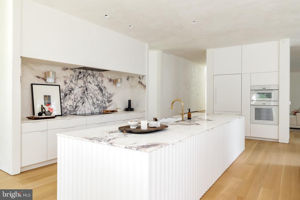 Modern white kitchen with a veined marble island, matching backsplash, brass faucet, and integrated wall ovens.