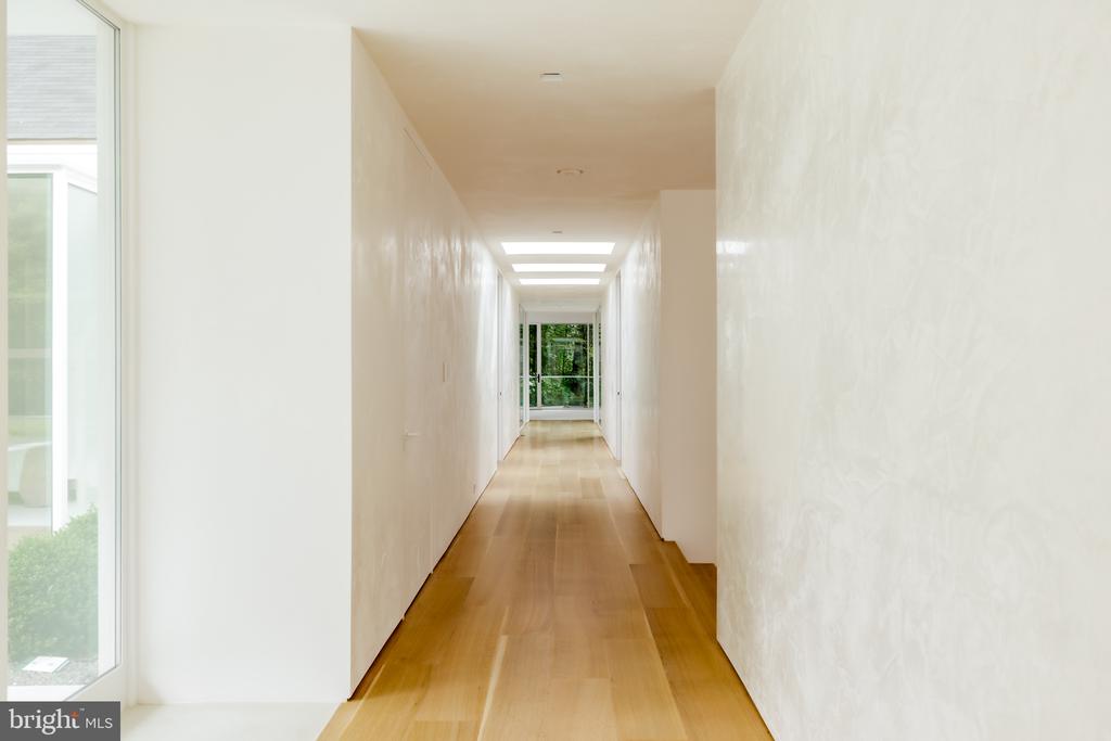 Empty hallway with light wood floors, ceiling skylights, and glass doors overlooking greenery.