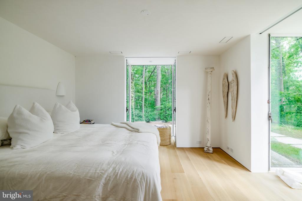 Modern bedroom with a large white bed and floor-to-ceiling windows overlooking trees.