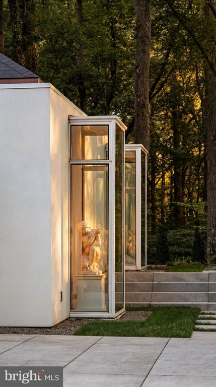 Modern white home with glass-fronted statue niches beside stone steps and mature trees.