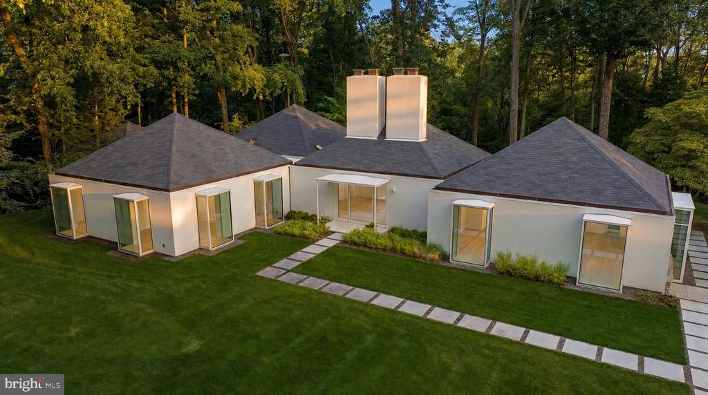 Modern white home with multiple hipped roof sections, a broad lawn, and a wooded backdrop.