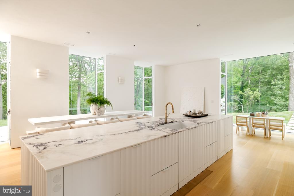 Modern kitchen with a long veined-marble island, brass faucet, and floor-to-ceiling windows overlooking trees.