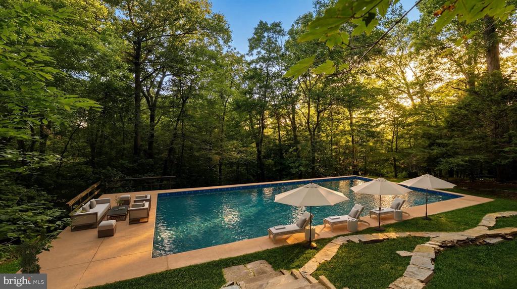 Rectangular backyard pool surrounded by forest, with patio seating and umbrellas at golden hour.