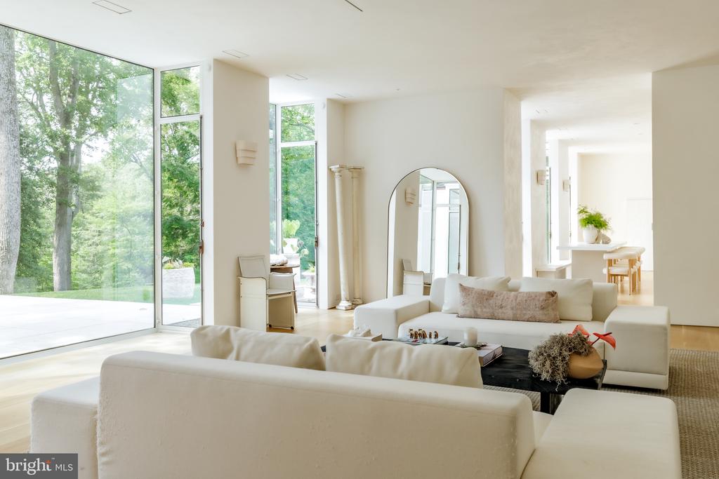 Bright living room with white sofas, floor-to-ceiling windows, and views of trees and a patio.