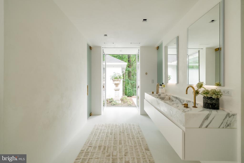 Bright bathroom with a marble double vanity and brass faucets, opening onto a garden.