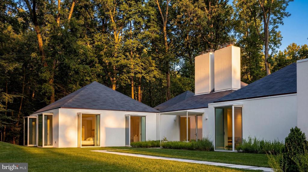 Modern white home with glass doors, tall chimneys, and a wooded backdrop in warm evening light.