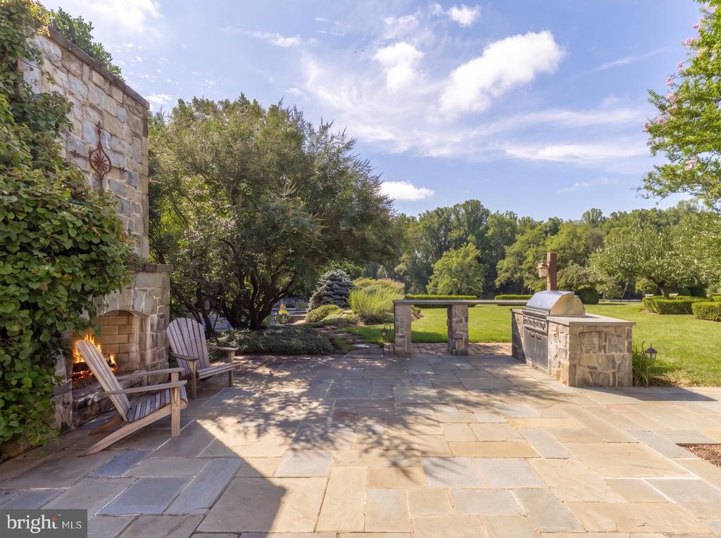 Stone patio with a fireplace, Adirondack chairs, and built-in grill overlooking a landscaped lawn.