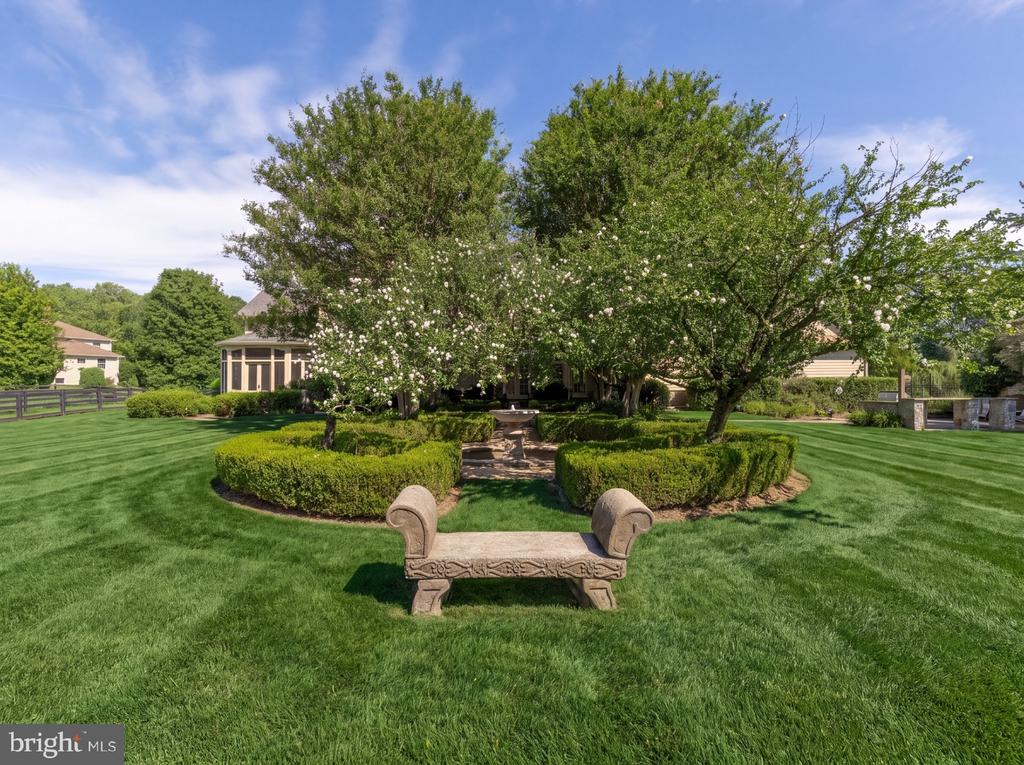 Landscaped yard with flowering trees, trimmed hedges, and a stone bench on the lawn.