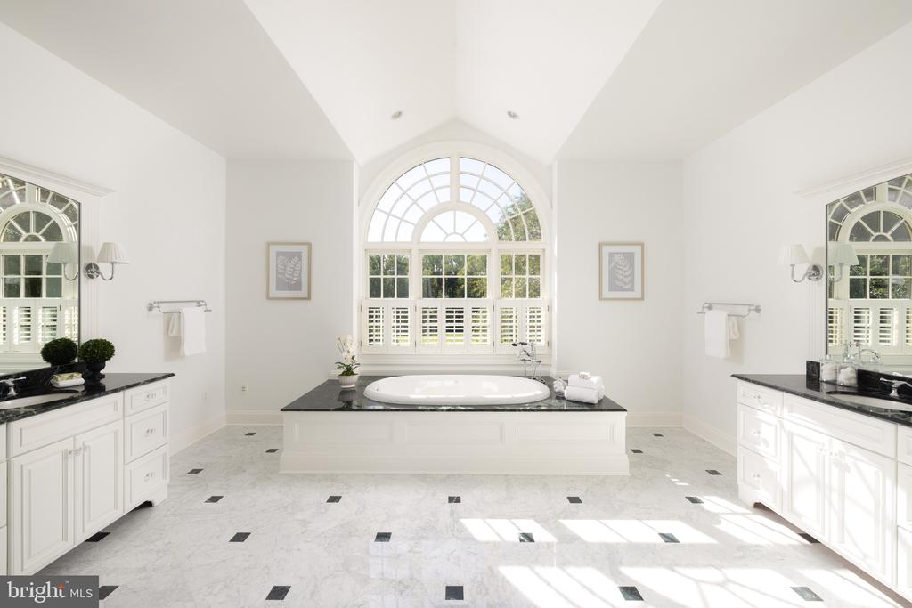 Primary bathroom with an oval tub beneath an arched window and white vanities with dark countertops.