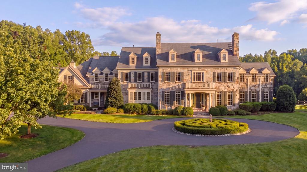 Large stone estate with dormers and chimneys, framed by a circular driveway and manicured landscaping.