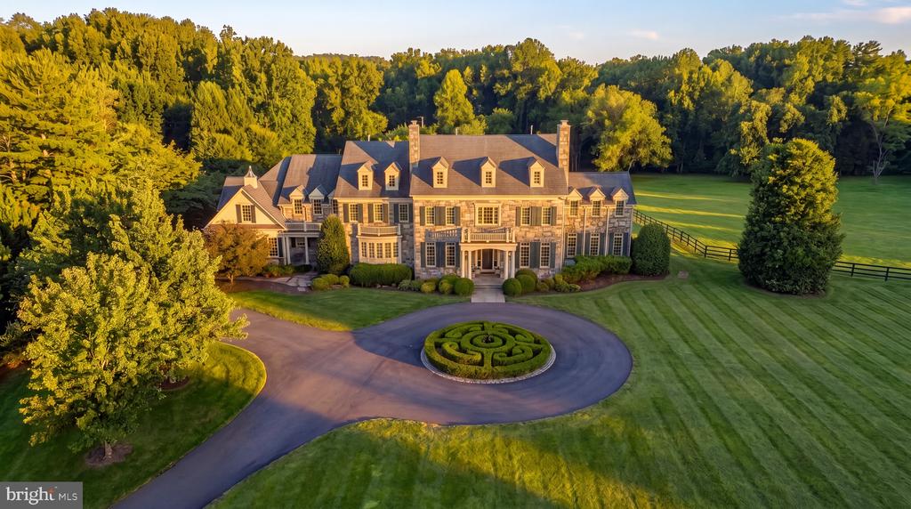 Aerial view of a large stone home with a circular driveway and clipped hedge maze at golden hour.